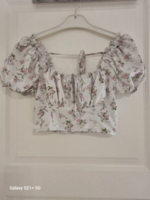 Blommig puffärm blus med knyt - Supersöt blommig blus med puffärmar och knyt framtill. Toppen är vit med små rosa och lila blommor, har croppad passform och resår vid axlarna för en trendig look. Perfekt för dig som vill ha en romantisk och somrig vibe.