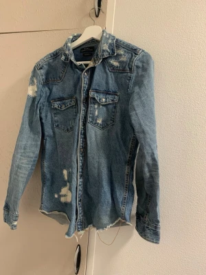 Ljusblå jeansskjorta med slitningar - Säljer en ljusblå jeansskjorta med slitna detaljer och råa kanter. Skjortan har två bröstfickor med lock och knappar framtill. Perfekt för en avslappnad och trendig look.