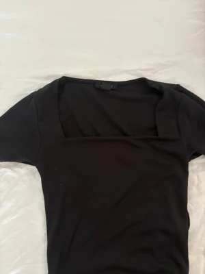 Svart topp med fyrkantig ringning - En stilren svart topp med korta ärmar och fyrkantig ringning. Toppen har en slim passform och är tillverkad i ett mjukt material som känns bekvämt mot huden. Perfekt att matcha med jeans eller kjol för en trendig look.
