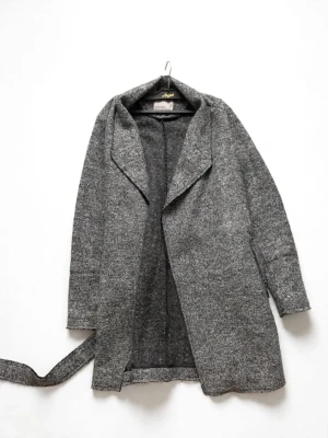 Grå lång kappa med skärp - Stilren grå kappa i ullblandning med avslappnad passform och bred krage. Kappan har ett matchande skärp i midjan och långa ärmar. Perfekt för kyliga dagar och enkel att styla till olika outfits.