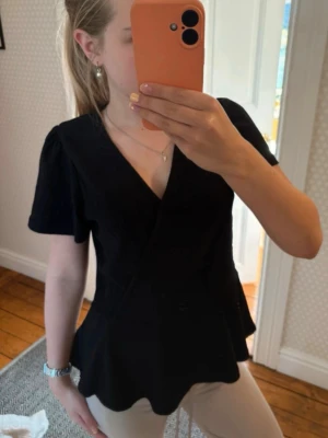 Svart omlottopp med v-ringning - Snygg svart topp med omlott-design och v-ringning. Toppen har korta ärmar och en lätt utsvängd nederkant. Perfekt att styla med både jeans och kjol för en trendig look.