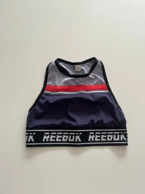 Sport-bh från Reebok  - Snygg sport-bh från Reebok med bred resår och logga runt om. Hög halsringning. Tillverkad i stretchigt funktionsmaterial som passar perfekt till träning. Tråden har gått upp lite på ett ställe (se sista bild), men annars i ett fint skick. Skulle säga att den är i storlek xs-s🤗