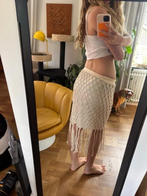 Beige strandkjol med fransar och nätmönster - Somrig beige kjol med nätmönster och långa fransar nedtill. Kjolen har en bred midjeresår och är perfekt för sommarens alla tillfällen. Materialet känns lätt och luftigt, och designen ger en bohemisk vibe. Perfekt att ha till stranded!