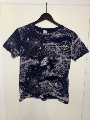 Mörkblå t-shirt med kartmönster Lindex - Mörkblå t-shirt från Lindex med unikt vitt kartmönster och kompassmotiv. Tillverkad i mjuk bomull och har en klassisk passform med rund hals och korta ärmar. Har ett litet hål i kragen, men inget man tänker på (se bild). Barnstorlek 146-152.