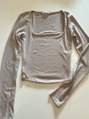 Beige långärmad topp med fyrkantig ringning - Snygg beige långärmad topp med en modern fyrkantig ringning. Toppen har en slim passform och är tillverkad i ett mjukt och stretchigt material som känns skönt mot huden. Perfekt att styla med jeans eller kjol för en trendig look.