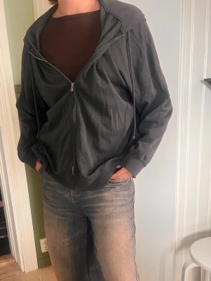 Mörkgrå zip hoodie med huva - Säljer en mörkgrå hoodie med dragkedja. Hoodien har långa ärmar, ribbade muddar och dragsko i huvan. Mjukt och bekvämt material.