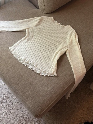 Vit ribbad topp från NA-KD - Säljer en vit långärmad topp från NA-KD i storlek 34. Toppen är ribbad med lätt vågiga kanter vid ärmslut och nederkant. Materialet känns lätt och stretchigt, perfekt för en stilren look.