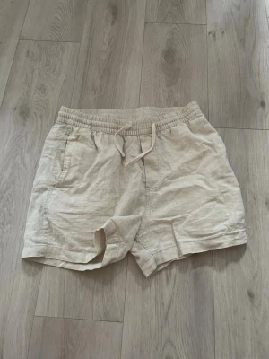 Beiga linneshorts från H&M - Säljer ett par beiga shorts från H&M i linnematerial. De har elastisk midja med snörning och två sidofickor. Regular fit och perfekt för varma dagar. Enkel och stilren design som passar till det mesta.