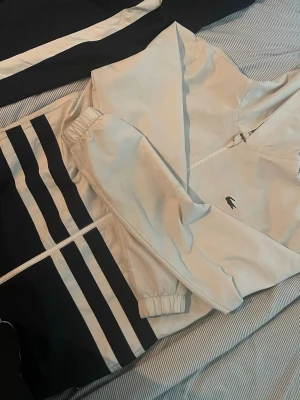 Vit och marinblå träningsjacka Lacoste - Snygg träningsjacka från Lacoste i vitt och marinblått med dragkedja och elastiska muddar. Jackan har klassiska ränder på ärmarna och liten krokodil-logga framtill. Perfekt för träning eller sportig stil.Lacoste tracksuit jacka M , passar perfekt som S också                                                                                   ny pris 3000kr  , säljs för 1799kr, ( billigare vid snabbaffär  ,fraktar samma dag ), vill få bort den så snabb så möjligt, inga märken inga skador precis som 