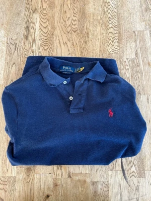 Marinblå piké från Polo Ralph Lauren - Klassisk marinblå pikétröja från Polo Ralph Lauren med röd broderad logga på bröstet. Slim fit-modell med krage och två knappar.