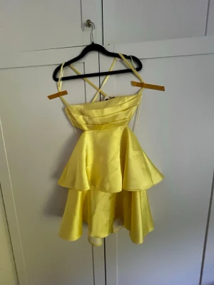 Gul kort klänning med volanger - Superfin gul kort klänning med volangkjol och smala axelband som korsas i ryggen. Klänningen har en omlottliknande överdel och är tillverkad i ett glansigt material som ger en lyxig känsla. Perfekt för dig som vill sticka ut med en färgglad look.