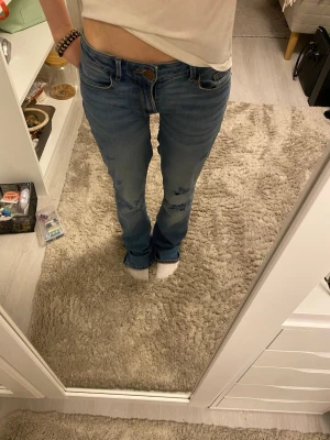 Blå slitna bootcut jeans - Säljer ett par blå bootcut jeans med slitningar på låren och knäna. Jeansen har klassisk femficksmodell och normal midja. Materialet är denim i bomull och passformen är avslappnad med lätt utsvängda ben. Omsydda för hand som du kan se på sista bilden