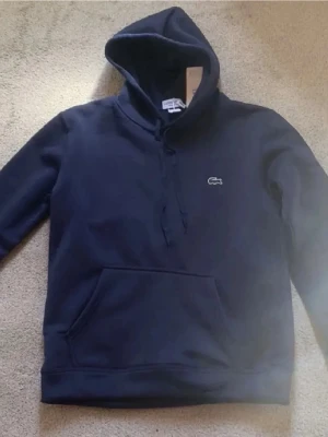Mörkblå hoodie från Lacoste - Snygg mörkblå hoodie från Lacoste med klassisk krokodillogo på bröstet. Hoodien har dragsko i huvan och en stor magficka framtill. Tillverkad i mjukt material som känns skönt mot huden. Perfekt för en avslappnad och stilren look.
