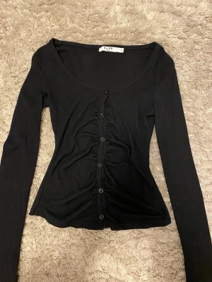 Svart ribbad topp från NA-KD - Snygg svart långärmad topp från NA-KD med ribbad struktur och knappar framtill. Toppen har en åtsittande passform och rynkade detaljer längs knappraden som ger en trendig look. Perfekt till jeans eller kjol.