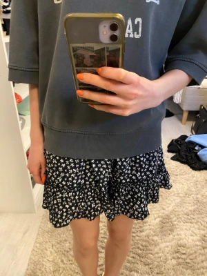 Svart blommig kortkjol med volang - Söt svart kortkjol med vitt blommönster och volangkant. Kjolen har en avslappnad passform och resår i midjan för extra komfort. Perfekt för dig som gillar en lekfull och trendig stil.