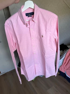 Rosa skjorta från Ralph Lauren - Säljer en klassisk rosa skjorta från Ralph Lauren med button-down krage och broderad logga på bröstet. Skjortan har långa ärmar och knappar framtill. Perfekt för dig som gillar stilrena plagg med en preppy vibe.