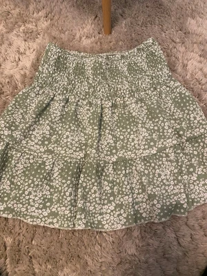 Grön blommig kortkjol Gina Tricot XS - Supersöt grön kortkjol från Gina Tricot i storlek XS. Kjolen har ett vitt blommigt mönster, smock i midjan och volangdetaljer. Perfekt för vår och sommar, lätt och luftig känsla.