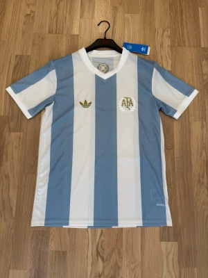 Argentina fotbollströja  - Säljer en retroinspirerad Argentina fotbollströja från Adidas i storlek S. Tröjan har klassiska ljusblå och vita vertikala ränder, v-ringad krage och guldfärgade detaljer med broderad logga och emblem. Tillverkad i lätt och ventilerande material.