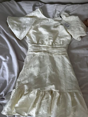 Vit kortklänning med volang - Söt vit kortklänning med strukturmönster och volang nedtill. Klänningen har korta, vida ärmar och markerad midja med fina detaljer. Perfekt för sommarens alla tillfällen.ny pris 800.