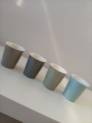 Fyra färgade porslinsmuggar - Säljer fyra stilrena porslinsmuggar i olika färger: beige, grå, ljusgrön och ljusblå. Muggarna har en enkel, rak form och vit insida. Perfekta för kaffe, te eller varm choklad. Snygga att ha framme i köket eller på kontoret.
