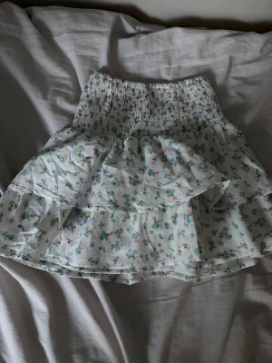 Vit blommig volangkjol - Supersöt kort kjol i vitt med små blommor i blått, rosa och grönt. Kjolen har bred smock i midjan och dubbla volanger som ger en luftig och lekfull känsla. Perfekt för varma dagar och enkel att matcha med en enkel topp.