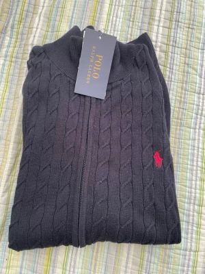 Mörkblå kabelstickad tröja Polo - Kabelstickad mörkblå tröja från Polo Ralph Lauren med dragkedja framtill och röd broderad logga på bröstet. Tröjan har hög krage och är tillverkad i mjukt stickat material. Perfekt för en stilren och klassisk look.