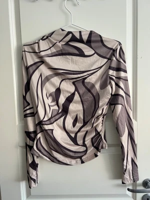 Beige mönstrad långärmad blus - Trendig beige blus med abstrakt svart och mörkbrunt mönster. Figurnära passform och långa ärmar. Perfekt för dig som vill sticka ut med en unik och modern look. Materialet känns mjukt och följsamt mot huden.