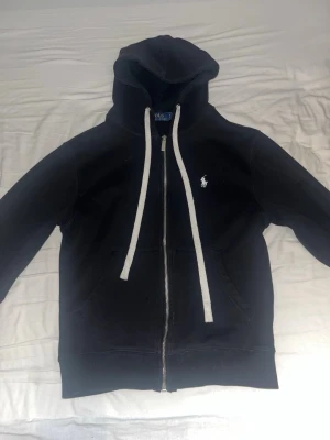 Svart hoodie från Polo Ralph Lauren - Svart hoodie med dragkedja från Polo Ralph Lauren. Klassisk design med vit dragsko, fickor framtill och den ikoniska vita logotypen broderad på bröstet. Tillverkad i mjukt bomullsmaterial. Storleken är ganska liten snarare S - XS.