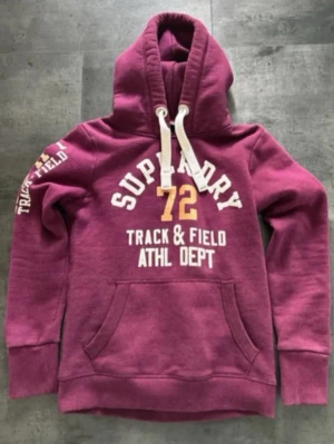 Vinröd hoodie från Superdry - Säljer en vinröd hoodie från Superdry med tryck i vitt och gult på bröstet och ärmen. Hoodien har stor känguruficka, tjocka vita dragsnören och mjukt material. Perfekt för en sportig och avslappnad stil.