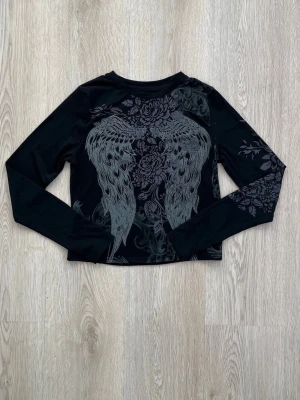 Svart långärmad topp med vingar - Cool svart långärmad topp med grått tryck av stora änglavingar och blommor på framsidan. Toppen har en croppad passform och är tillverkad i ett stretchigt syntetmaterial. Perfekt för dig som gillar edgy detaljer och unika prints.