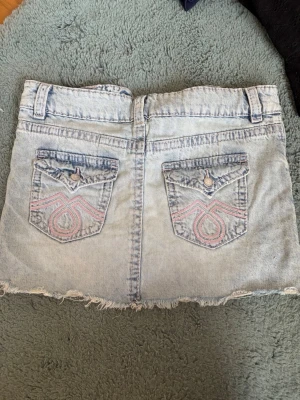 Ljus jeanskjol med broderade fickor - Säljer en ljusblå jeanskjol med rå kant och två bakfickor med rosa broderi och knappdetaljer. Kjolen har bälteshällor och en klassisk denimlook, perfekt för dig som gillar en avslappnad stil.
