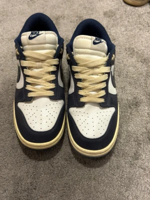 Nike Dunk Low blå och vita sneakers - Snygga Nike Dunk Low sneakers i marinblått och vitt läder med beige snören. Klassisk siluett med perforerad tåbox och vit sula. Perfekta för dig som gillar retrostil och streetwear. Från 🇨🇳