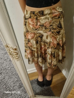 Mönstrad kjol från Ibn Al Arab - Säljer en unik kjol från Ibn Al Arab med paisley- och blommönster i beige, brunt och inslag av rosa. Kjolen har en dold dragkedja i sidan och är tillverkad i ett glansigt material som känns lyxigt. Perfekt för dig som vill sticka ut med en bohemisk vibe.