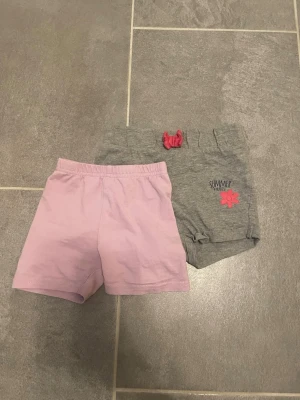 Två par mjuka shorts i rosa och grå - Säljer två par bekväma shorts, ett par i ljusrosa och ett par i grått med rosa rosett och blomdetalj samt texten 'SUMMER'. Båda har elastisk midja och är perfekta för varma dagar. Tillverkade i mjuk bomull för skön känsla.