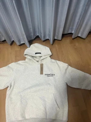 Ljusgrå Essentials Fear of God hoodie - Snygg ljusgrå hoodie från Essentials Fear of God med svart logga på bröstet. Klassisk modell med huva och känguruficka framtill. Perfekt för en avslappnad och trendig stil. Mjuk bomullsblandning som känns skön mot huden.