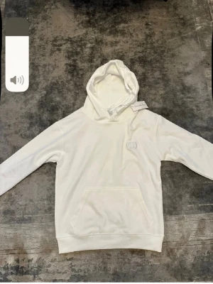 Vit hoodie med broderad logga - Snygg vit hoodie med huva och känguruficka framtill. Diskret broderad logga på bröstet och ribbade muddar vid ärmslut och nederkant. Tillverkad i mjukt material som känns skönt mot huden. Perfekt för en avslappnad och stilren look.
