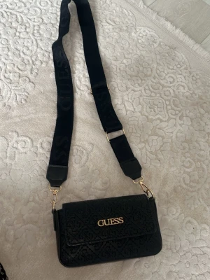 Svart axelväska från Guess - Snygg svart axelväska från Guess med präglat mönster och guldfärgad logga framtill. Väskan har en bred, justerbar axelrem med Guess-tryck och guldfärgade metalldetaljer. Perfekt storlek för det viktigaste och ger en trendig touch till din outfit.