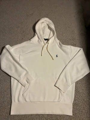 Vit hoodie från Polo Ralph Lauren - Snygg vit hoodie från Polo Ralph Lauren med klassisk broderad logga på bröstet. Hoodien har dragsko i huvan och en stor magficka. Tillverkad i mjukt material som känns skönt mot huden. Perfekt för en clean och stilren look. Har en liten liten fläck vid fickan men syns absolut ingenting när man använder den!!! Storlek M priset ej hugget, vid snabb affär kan priset sänkas