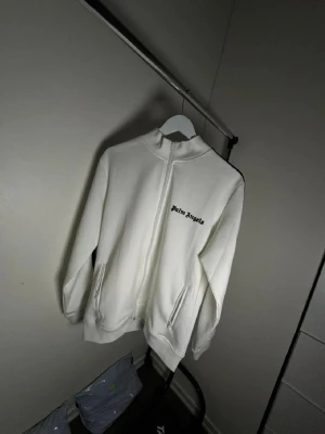 Vit zip-hoodie från Palm Angels - Snygg vit hoodie med dragkedja från Palm Angels. Svarta ränder längs ärmarna och logga på bröstet. Hög krage och två fickor framtill. Perfekt för en clean och sportig look.