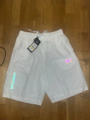 Vita shorts från Under Armour - Snygga vita shorts från Under Armour med resårmidja och snörning. Reflekterande logga och textdetaljer på framsidan samt reflekterande ränder på baksidan. Perfekta för träning eller chill. Två sidofickor och luftig passform.