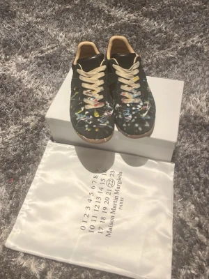 Svarta sneakers med färgstänk - Unika svarta sneakers från Maison Margiela med färgstänk i flera nyanser och beige detaljer. Skorna har vita snören och en klassisk låg siluett. Tillverkade i läder med en cool konstnärlig vibe. Perfekta för dig som vill sticka ut med din stil.
