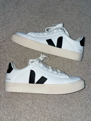 Vita Veja Campo sneakers med svart detalj - Snygga vita sneakers från Veja med klassisk svart V-logga på sidan och vit sula. Skorna har lågt skaft, vita skosnören och stilren design som passar till det mesta. Tillverkade i slätt läder med bekväm innersula. Aldrig använd.