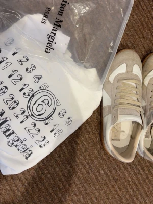 Maison Margiela vita och beige sneakers - Maison Margiela sneakers i vitt och beige med klassisk retrodesign. Ovandelen är i mocka och läder, med vita snören och gummisula. Snygg kontrast mellan material och färger, perfekt för en clean och stilren look.