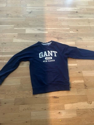 Mörkblå sweatshirt från GANT XS - Säljer en mörkblå sweatshirt från GANT i storlek XS. Tröjan har rund halsringning och vit text med 'GANT 1949 NEW HAVEN' på bröstet. Tillverkad i mjukt bomullsmaterial och har klassisk college-look. Perfekt för dig som gillar stilrena och bekväma plagg.