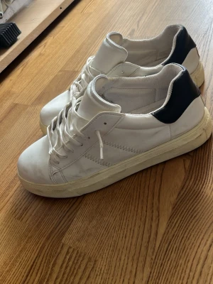 Vita sneakers med svart detalj - Snygga vita sneakers med svart hälparti och vita snören. Skorna har en klassisk låg profil och är tillverkade i slätt lädermaterial. Perfekta för en clean och stilren look till vardagsoutfiten.