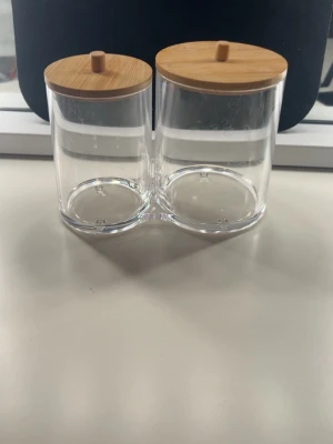Tvådelad burk med bambulock - Säljer en genomskinlig tvådelad burk i plast med runda lock i ljust bambuträ. Perfekt för förvaring av småsaker, som tops och pads. Burkarna sitter ihop i botten och locken har små knoppar för enkel öppning.