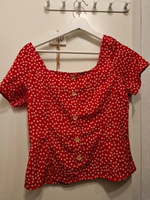 Röd blommig blus med knappar - Söt röd blus med små vita blommor och träknappar framtill. Blusen har korta ärmar och en något vid, feminin passform. Perfekt för dig som gillar färg och mönster!
