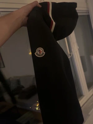 Svart Moncler jacka storlek 3 - MER BILDER SKICKAS VID INTRESSE Snygg svart jacka från Moncler med klassisk logotyp framtill och stilrena röda och vita ränder upptill. Perfekt sommarjacka för att hålla värmen och samtidigt se trendig ut. Passar dig som gillar exklusiva märken och enkel design.
