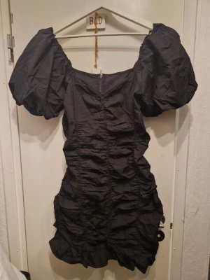 Svart kort klänning med puffärmar - Trendig svart kort klänning med stora puffärmar och rynkade detaljer längs hela klänningen. Klänningen har en figurnära passform och dragkedja i ryggen. Perfekt för dig som vill sticka ut med en cool och modern look. Hittar ingen storlek, men jag har m och den passar mig. Köpt på gina, men bara hängt i garderoben mestadels.