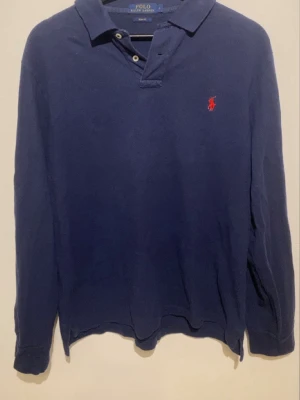 Marinblå långärmad skjorta Polo - Marinblå långärmad tröja från Polo Ralph Lauren med klassisk krage och röd broderad logga på bröstet. Tillverkad i mjuk bomull för en avslappnad men stilren look. Den är slim fit i storlek L, och har inga defekter överhuvudtaget. Priset är inte hugget i sten, släng iväg ett dm om du har några frågor 😁